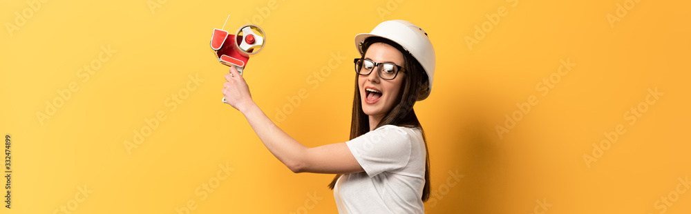 panoramic shot of shocked handywoman holding tape dispenser on yellow background panoramic shot of shocked handywoman holding tape dispenser on yellow background