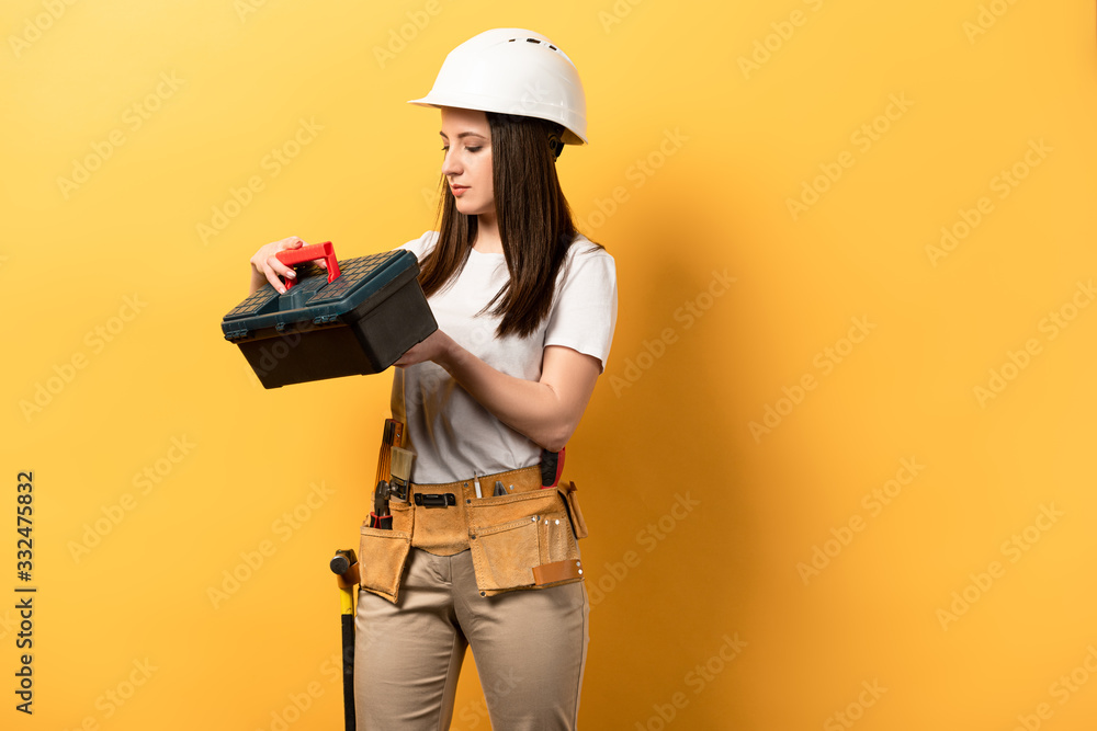 attractive handywoman in helmet holding toolbox on yellow background
