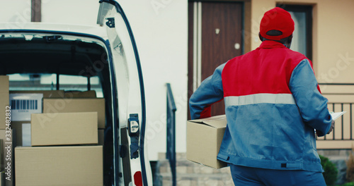 Attractive young African American mailman in red costume and cap taking out percel from a van and walking to the house to deliver it. Outdoors.