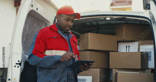 African American young mailman in red costume and cap counting the mail boxes in a van with tablet device in hands. Outdoors.