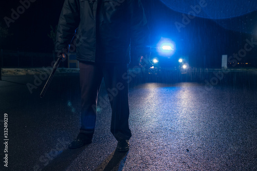 Police man with mask and nightstick and walkie talkie in hand watching and patrol car with sirens and blue lights on the curfew in the streets during the state of alarm in the covid 19 coronavirus. photo