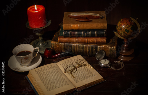 An old desk with a pile of vintage brown leather books, an open book with eye glasses, a cup of coffee, a red candle with flame, a vintage pocket clock, earth globe.