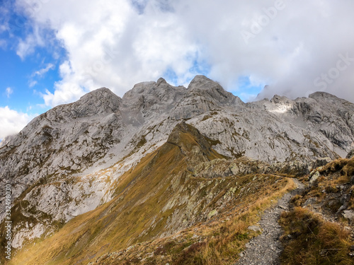 A pathway leading to high peaks in Italian Alps. Sharp slopes on both sides of the valley. Hard to reach mountain peaks. There are many mountain ranges in the back. Serenity and peace. Autumn vibes