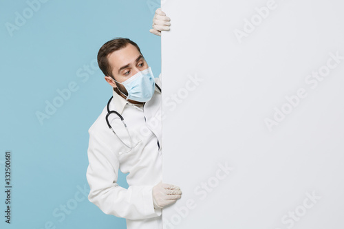 Male doctor man in gown face mask gloves isolated on blue background. Epidemic pandemic coronavirus 2019-ncov sars covid-19 flu virus. Hold big white empty blank billboard with place for text image. photo