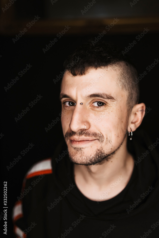 Confident adult male with piercing and mohawk smiling and looking at camera in dark room Confident adult male with piercing and mohawk smiling and looking at camera in dark room
