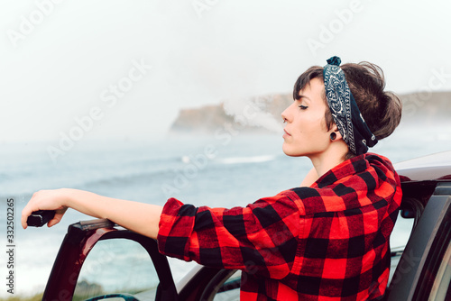 Side view of extraordinary woman with black short hair and bandana on head and piercing in red plaid shirt leaning on car and looking away at seaside photo