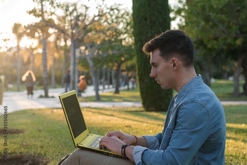 Side view of smart concentrated man with backpack studying at laptop sitting in park grass with crossed legs in sunny day Side view of smart concentrated man with backpack studying at laptop sitting in park grass with crossed legs in sunny day