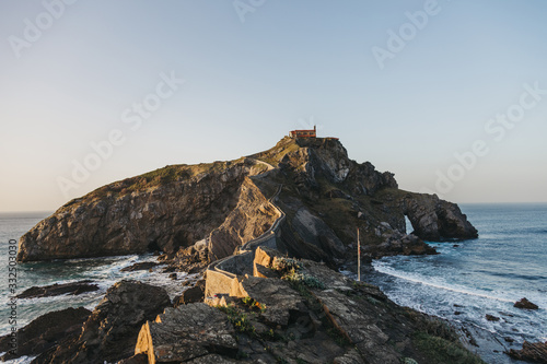 Paving stone way leading along stone bridge and ridge of rocky hill to lonely country house on island Gaztelugatxe surrounded by tranquil sea water with white foam waves