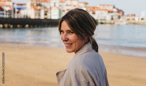 Happy blond female in casual clothes looking at camera laughing while standing on pier and touching hair against blurred urban environment of resort town in Spain