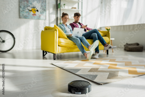 selective focus of robotic vacuum cleaner washing carpet near man watching movie and woman using laptop in living room
