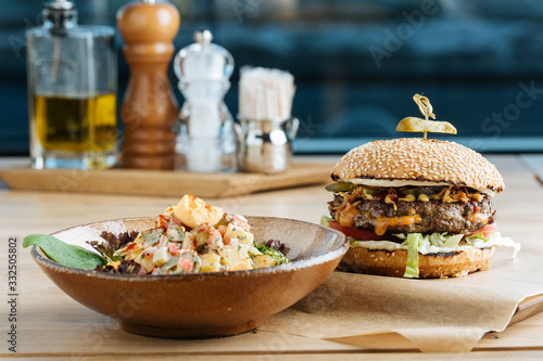 Big hamburger on paper with cutlet cheese and vegetables served with bowl of colorful salad in modern restaurant