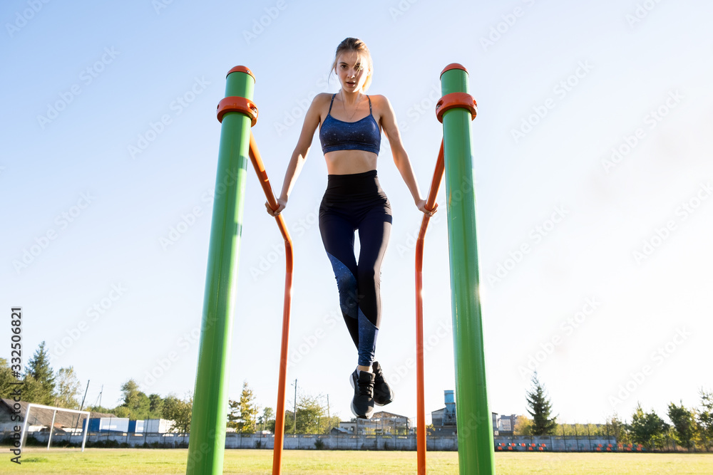Fototapeta premium Young sportive athlete woman doing fitness exercise on metal bars outdoors at stadium court.