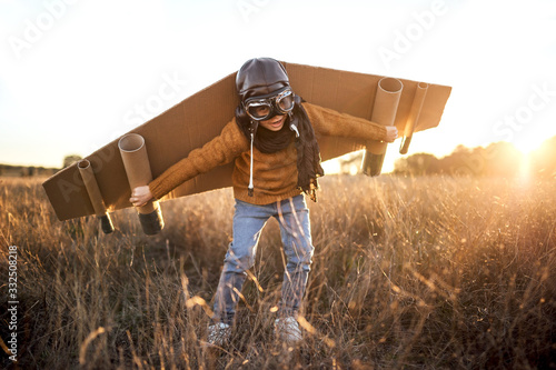 Happy kid in goggles and cardboard wings raising hands during game on field in backlit photo