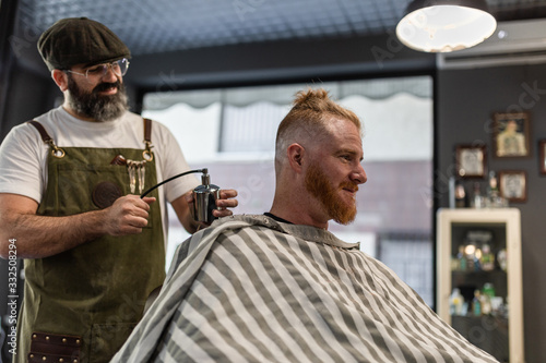 Barber applying fixative spray while making trendy hairstyle for confident relaxed man in cozy beauty salon photo