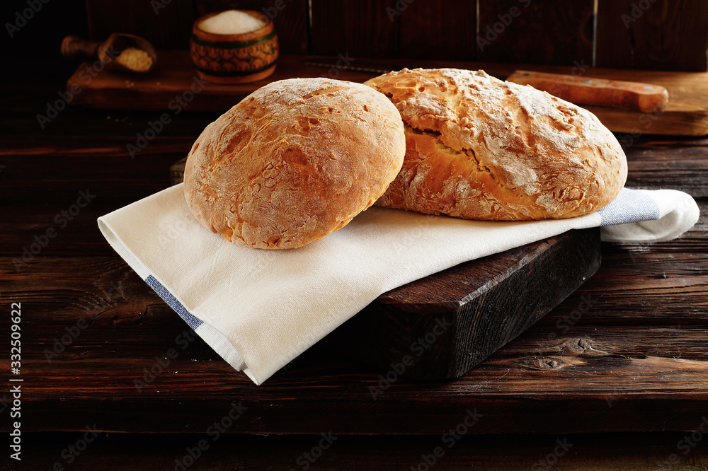 Two loaves of wheat bread made at home lie on a white, linen napkin and a wooden Board. In the background, a salt shaker, a sesame scoop, and a bread knife. Fresh baked goods , close -up.
