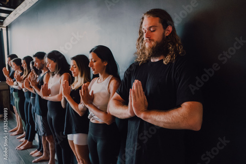 Group of happy young sporty people in activewear with eyes closed relaxing and deep breathing while standing with namaste in tadasana and meditating beside black wall in modern yoga studio photo