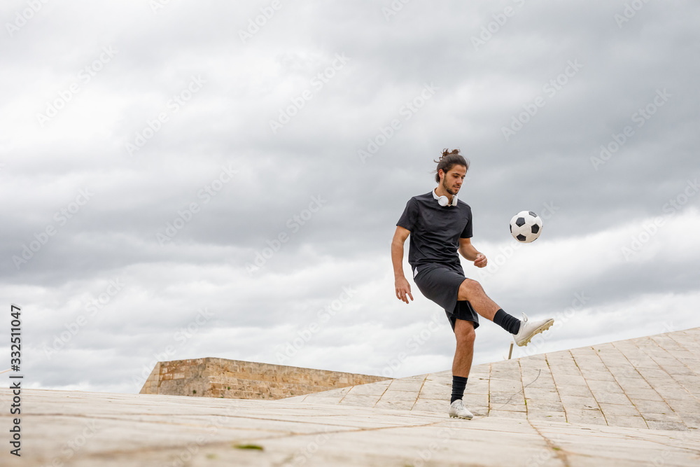 Young sportive male in black t shirt and shorts kicking soccer ball while training on paved street with cloudy sky in background Young sportive male in black t shirt and shorts kicking soccer ball while training on paved street with cloudy sky in background