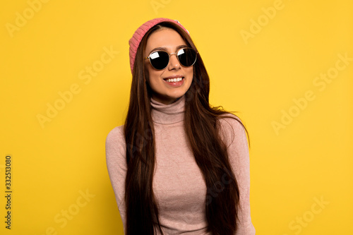 Modern stylish hipster female with long dark hair wearing pink cap and round sunglasses posing with happy smile over yellow background