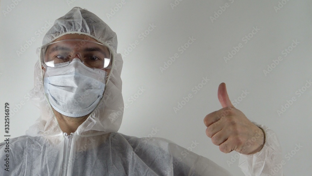 A man in protective clothing. On the face are glasses and a mask. Holds a thumbs up. Portrait of a man. The fight against the virus. Light background A man in protective clothing. On the face are glasses and a mask. Holds a thumbs up. Portrait of a man. The fight against the virus. Light background