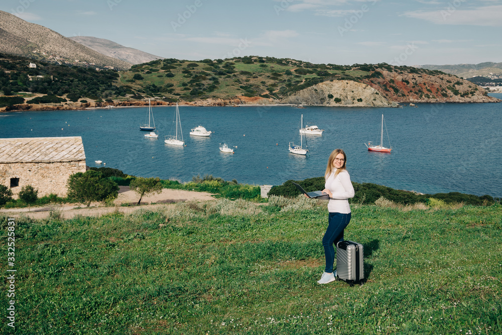 Pretty blonde female freelancer working at laptop at the hill with panoramic view to the sea and boats