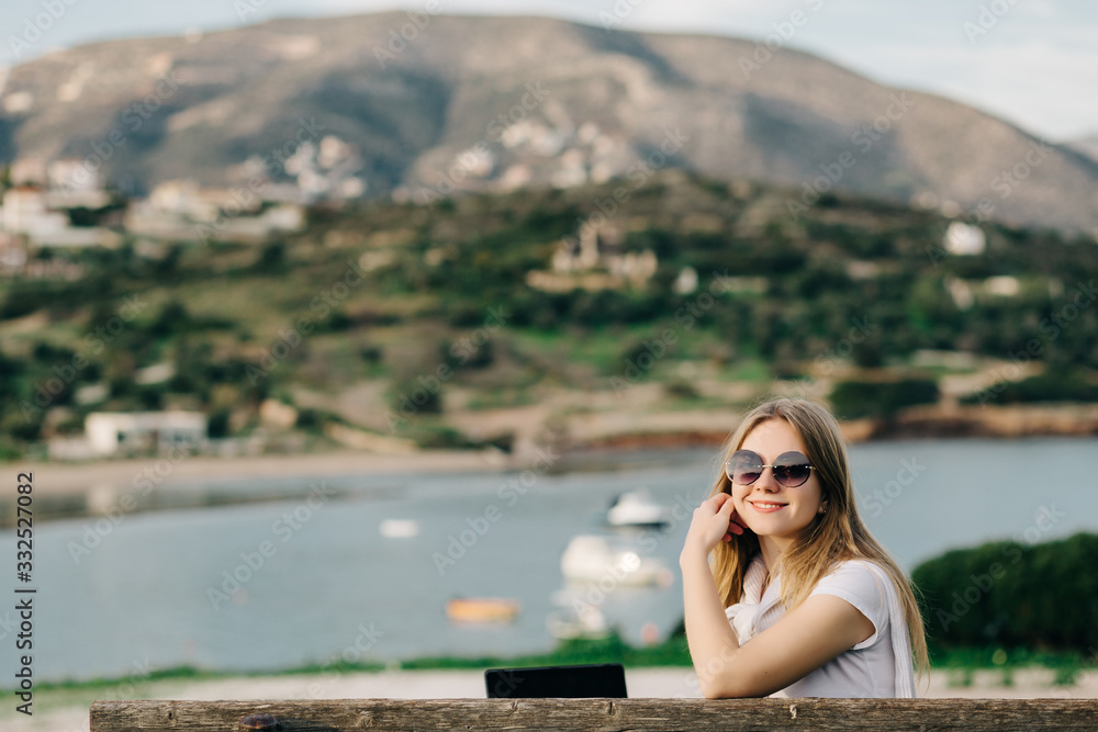 Happy young girl in glasses sits on a bench on a hill by the sea