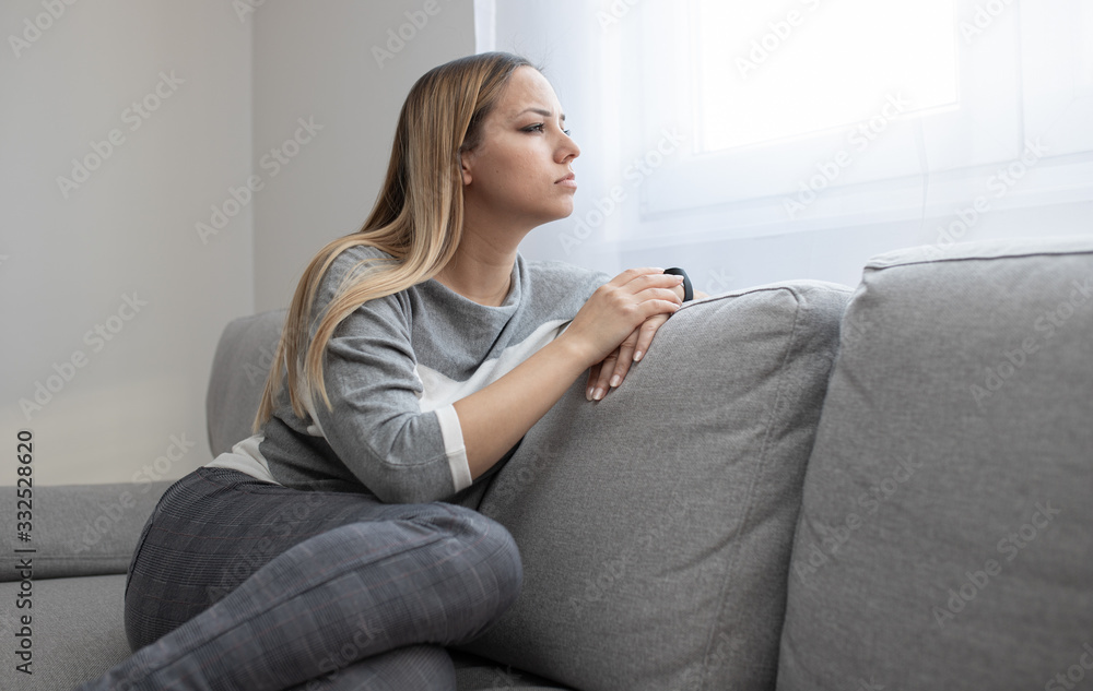 Sad and depressed woman sitting on sofa and looking at distance. Woman bored at home.