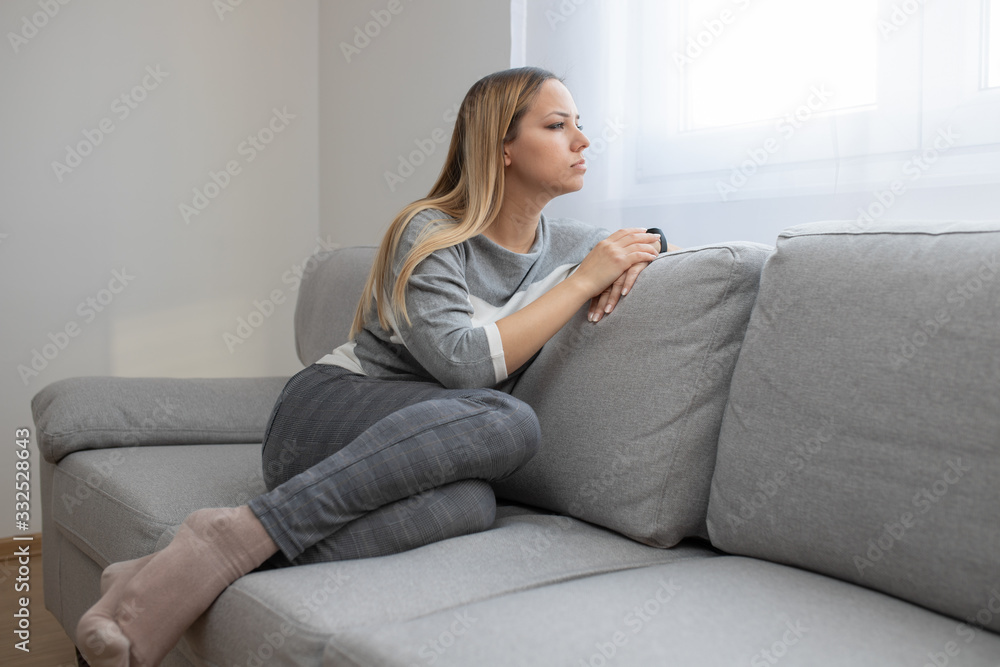 Sad and depressed woman sitting on sofa and looking at distance. Woman bored at home.