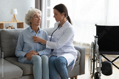 Young woman doctor visit senior disabled grandmother at home listen to heart rate use stethoscope, attentive female nurse take care of sick mature grandma on wheelchair, elderly healthcare concept