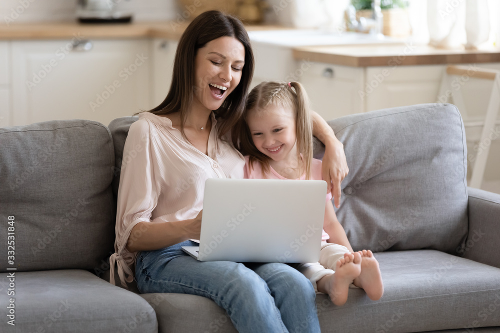 Overjoyed young mother and cute little daughter have fun relax at home together watch funny video cartoon on laptop, smiling mom and small girl child spend leisure time using computer playing game
