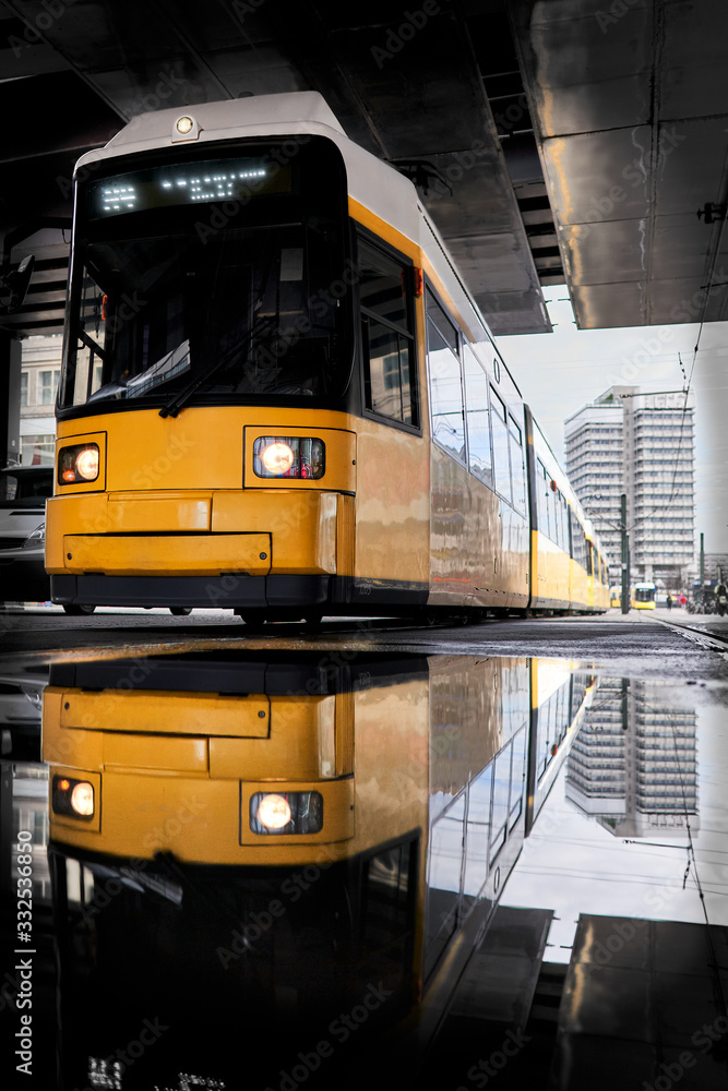 yellow tram is reflected in a street puddle on alexander Platz in the centre of Berlin, the train goes through a dark bridge, in the background there is a white blurred high-rise building