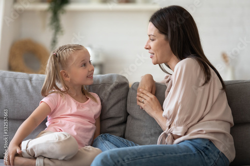 Happy young mother and little preschooler daughter sit on couch in living room talking and chatting, smiling small girl child enjoy leisure time with mom speak communicate spend time together