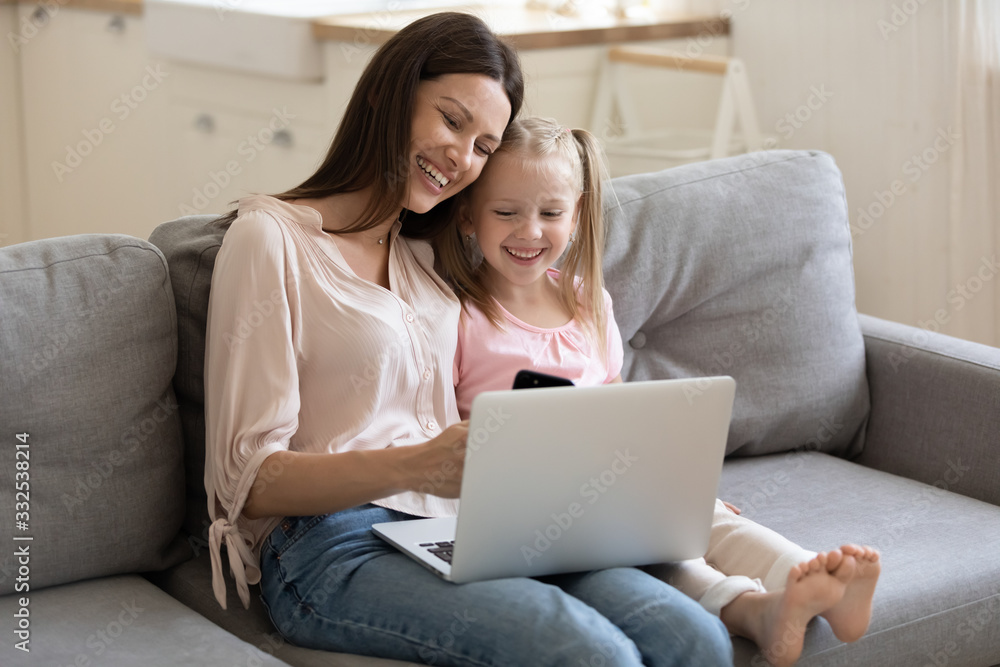 Naklejka premium Happy young mom and small smiling daughter sit rest on couch at home watching funny video on laptop, overjoyed mother and cute little girl child have fun using devices enjoy weekend together