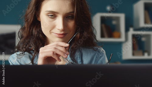 Headshot of young cheery woman holding spectacles looking at computer screen, front view