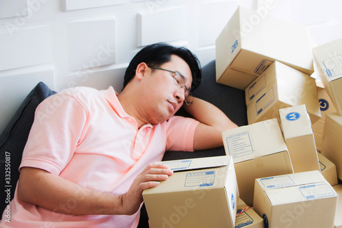 A middle-aged Asian male worker is taking a nap beside a pile of mass packed boxes. After his shop received a lot of orders from online stores because of the Covid-19 virus.