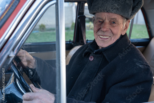 Happy smiling old man in black jacket driving retro car, holds her hands on wheel and looking at the camera