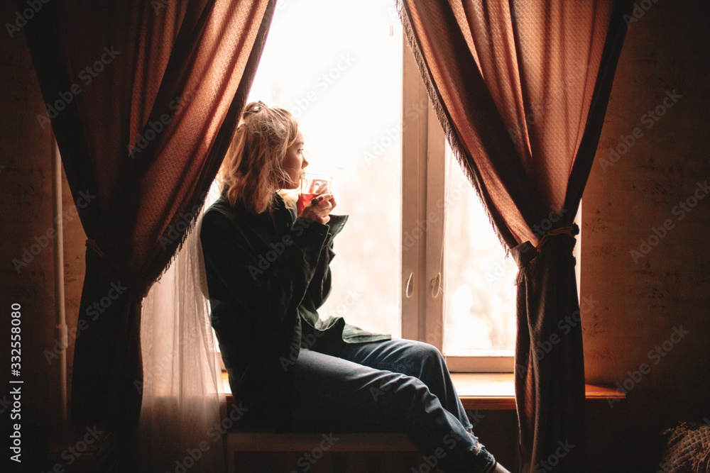 Obraz premium Young thoughtful woman drinking tea and looking through window while sitting on windowsill at home