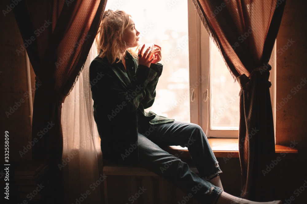 Young thoughtful woman drinking tea and looking through window while sitting on windowsill at home