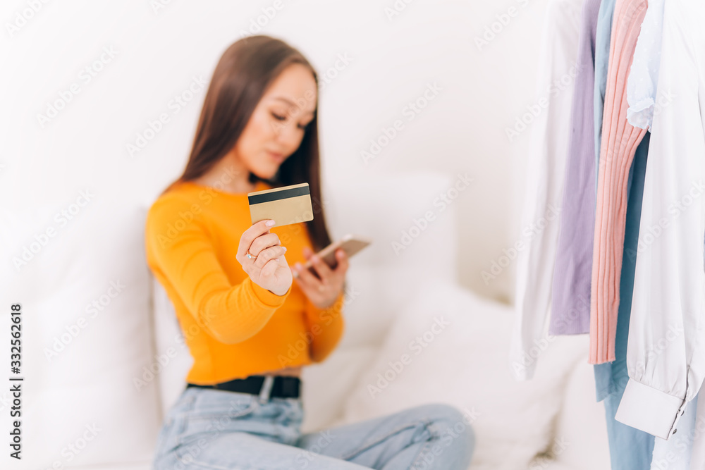 A young girl sitting on a sofa with a credit card and a phone, pays for a purchase, pays bills, online payment in an orange sweater A young girl sitting on a sofa with a credit card and a phone, pays for a purchase, pays bills, online payment in an orange sweater