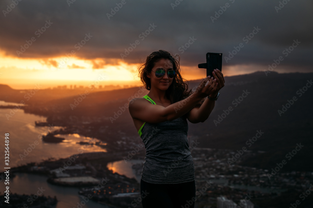 Girl on a sunset hike with ocean and city view is taking a selfie on a smartphone