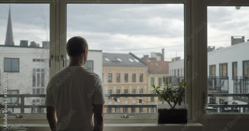 Wide static back cinema camera shot of frustrated Norwegian male in white t-shirt isolated alone in Corona virus quarantine looking out window from dark apartment with city background, in Oslo Norway. Wide static back cinema camera shot of frustrated Norwegian male in white t-shirt isolated alone in Corona virus quarantine looking out window from dark apartment with city background, in Oslo Norway.
