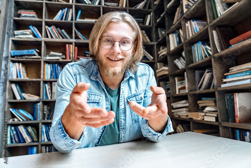 Фотография Young happy smiling student man hipster blogger vlogger glasses have online interview skype call shoot self presentation video log explain tell story chat look at camera home library point at camera