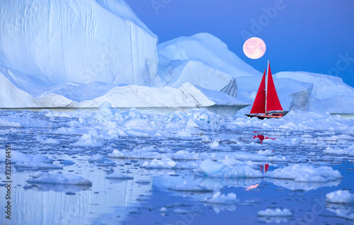 Sail boat with red sails cruising among ice bergs after sunset in front of a full moon. Disko Bay, Greenland.