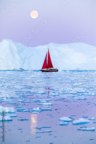 Sail boat with red sails cruising among ice bergs after sunset in front of the full moon rising. Disko Bay, Greenland.
