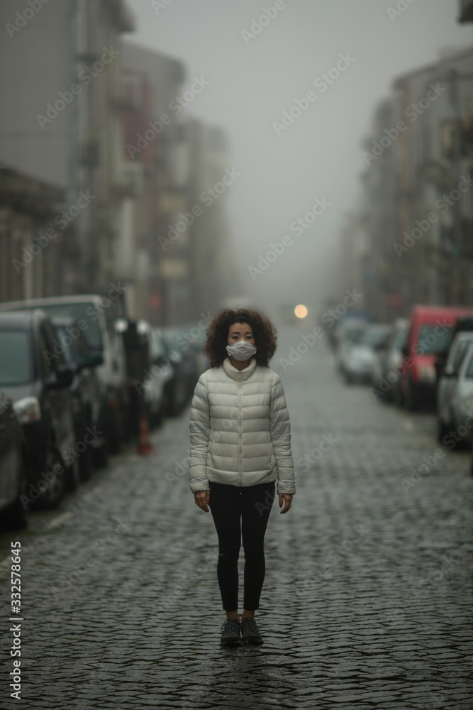 Young mixed-race asian woman in antiviral mask stands in the street in cloudy weather. Pandemic. COVID-19, Coronovirus.