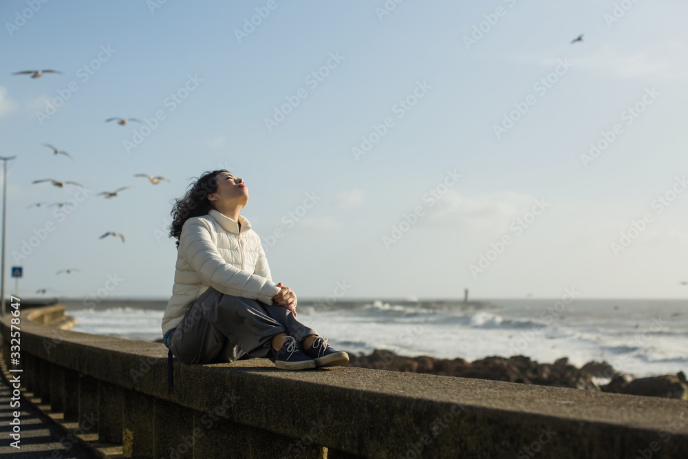 Beautiful mixed-race asian woman sitting of sea promenade. Seagulls fly over the foaming surf.