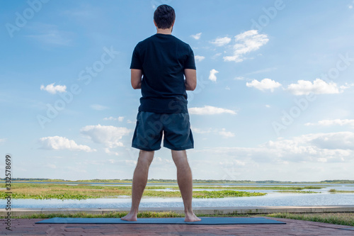 Man wearing a black shirt and dark green shorts stands looking outward over the sea in attempt to correct his bodies posture in front of a bright blue sky with gorgeous white clouds behind his body