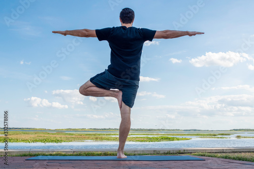 Man wearing a black shirt and dark green shorts stands straight up on one leg in vriksasana correcting his bodies posture in front of a bright blue sky with gorgeous white clouds behind his body. photo