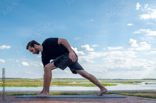 Man wearing a black shirt and dark green shorts bends over in a triangle pose correcting his bodies posture in front of a bright blue sky with gorgeous white clouds behind his body.