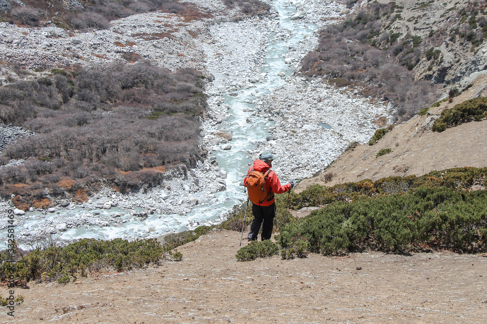 Person in orange windbreaker walks on footpath during sunny day in Himalayas on the way to Everest base camp. Imja Khola river flows in valley in the background. Theme of trekking in Nepal.