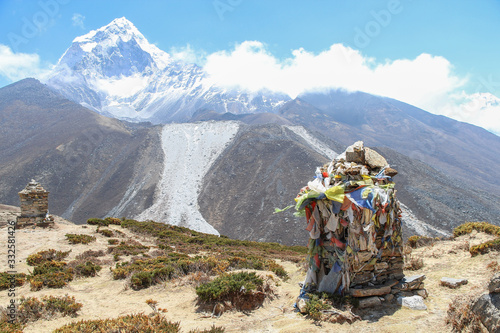 Monuments to the dead climbers covered with buddhist prayer flags in the foreground. Ama Dablam mountain peak and clouds in the background. Footpath to Everest base camp in Himalayas in Nepal. 